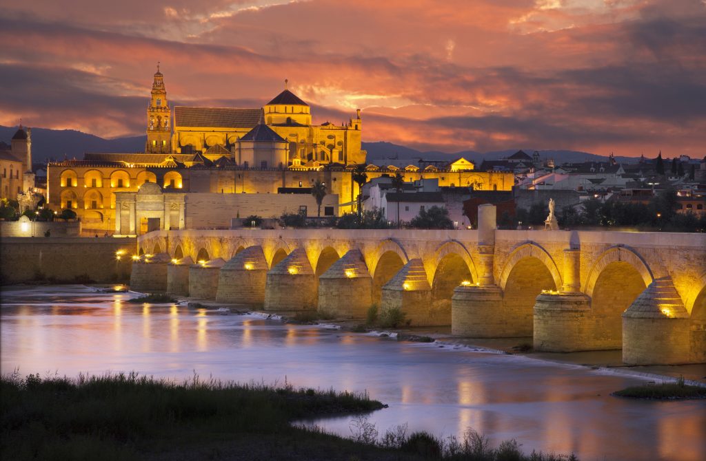 Cordoba - The Roman bridge and gate with the Cathedral in the background at evening dusk - Accent On Travel